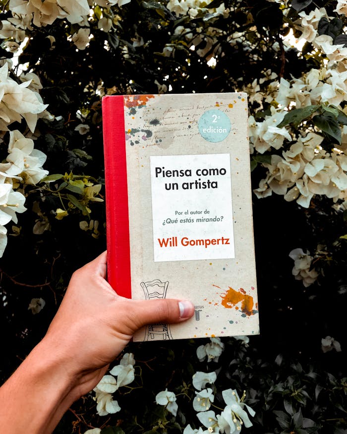 A book titled Piensa como un artista held in front of white flowers.